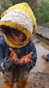 child-looking-at-his-muddy-hands-whilst-stood-in-a-muddy-area-wearing-raincoat-and-balaclava-style-knitted-hat.